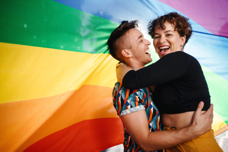 Happy,Queer,Couple,Standing,Against,A,Rainbow,Pride,Flag.,Young